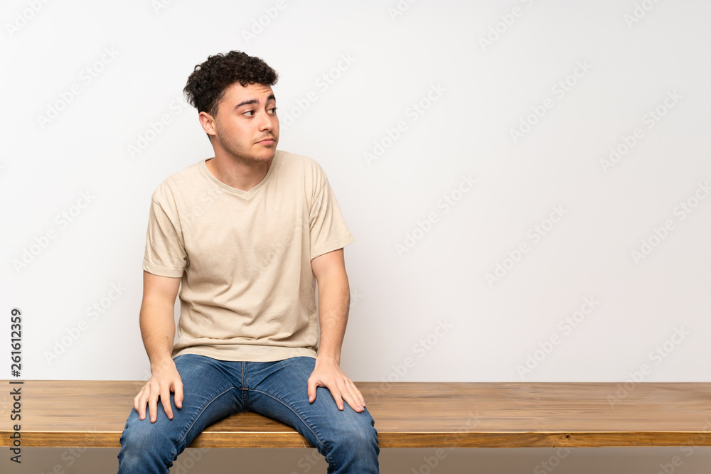 Young man sitting on table making doubts gesture looking side Young man sitting on table making doubts gesture looking side
