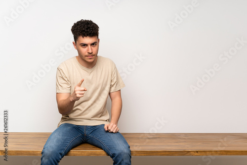 Young man sitting on table frustrated and pointing to the front