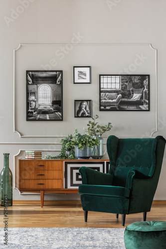Emerald green armchair in elegant living room with black and white photos on grey wall and retro cabinet