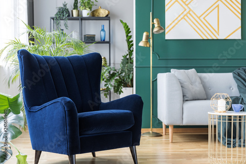 Blue armchair next to grey scandinavian sofa in tropical inspired interior with green and gold colors photo