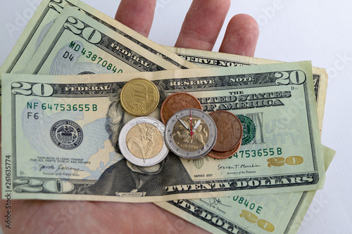 time for currency exchange: 2 euro coin with hour hands on a pile of dollars, several different eurocents nearby, in the person’s hand, short focus, close photo