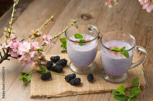 Fresh fruit smoothie blackberry milkshake, purple drink on wood table with sprigs of mint, fresh blackberries, and cherry blossoms. Springtime healthy shake recipe in beautiful setting.