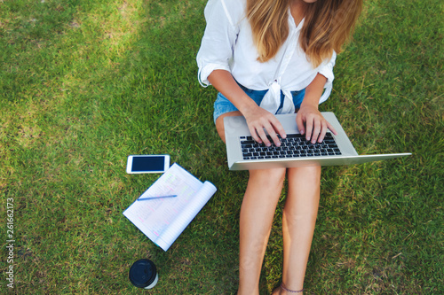 Top view of woman sitting in park on the green grass with laptop, notebook and smartphone, hands on keyboard.