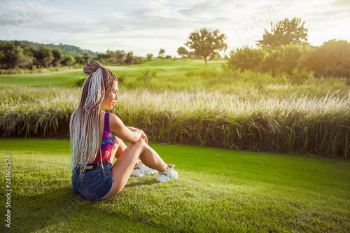 Girl with African braids sitting on the lawn and watching the sunset. copy space