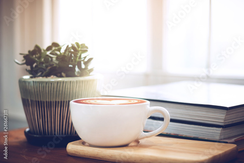Closeup of latte art coffee on wooden table with book and small tree in ceramic vase background, vintage color tone