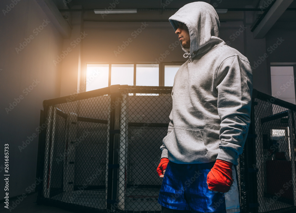 Portrait of young male athlete dressed in grey hoodie, with hood down on eyes, engaging in boxing activity to increase the fortitude, posing in gym in red hand bandages. Sport, hobby, martial arts. Portrait of young male athlete dressed in grey hoodie, with hood down on eyes, engaging in boxing activity to increase the fortitude, posing in gym in red hand bandages. Sport, hobby, martial arts.