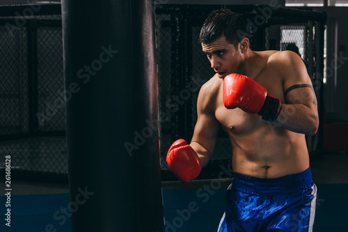 Handsome determinated bare chested male boxer in red gloves getting prepared for big fight, doing cardio boxing workout with punching bag in empty dark gym. Sport, challenge victory, workout. © alfa27