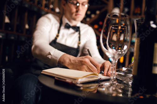 Blurred photo of male cavist making notes at his notepad about wine features and origin in wine shop, focus on clean empty wine glass
