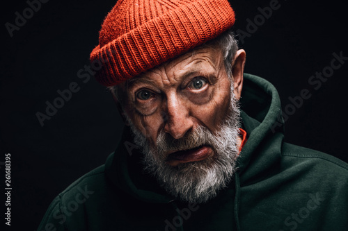 Elderly man indoor studio portrait wearing orange hat, having white beard and wrinkled weathered face, looking at camera with crazy confused expression. Mental Aging Disorders concept © alfa27