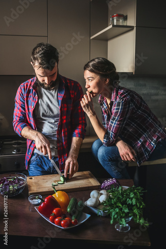 Beautiful young couple in kitchen at home while cooking healthy food. Man is cuts salad. Woman sits on table eating a cucumber and cunningly looks at her husband. Scene from family life.