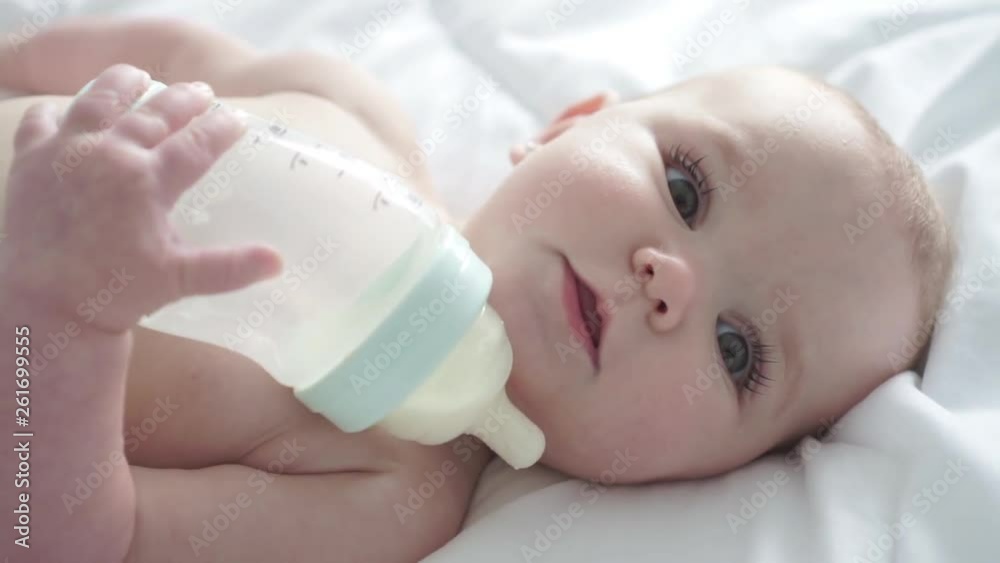 Pretty baby girl drinks milk from bottle lying on bed. Child weared diaper in nursery room. Pretty baby girl drinks milk from bottle lying on bed. Child weared diaper in nursery room.