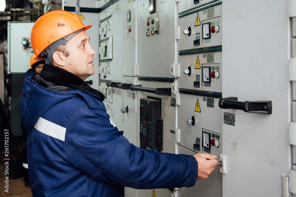 A man in overalls wearing a protective helmet is standing by an electrical shield. A man in overalls wearing a protective helmet is standing by an electrical shield.