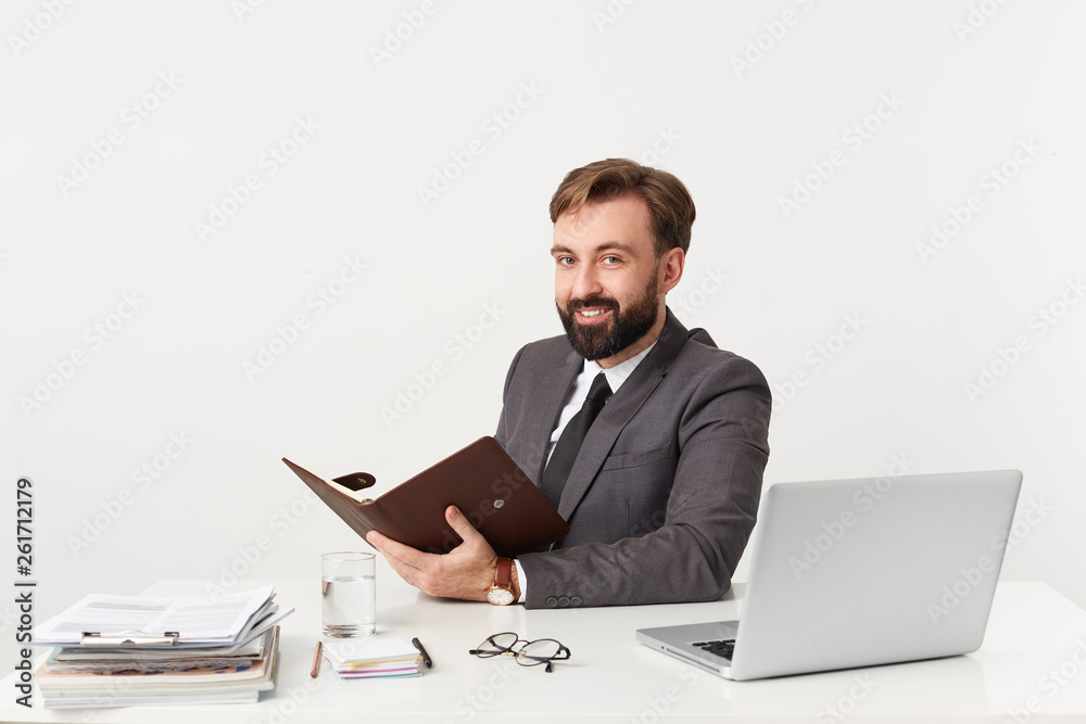 Attractive smiling successful businessman, top manager with beard holding his diary sitting at desktop in office, looking camera, dressed in a expensive suit with a tie, has a good watch Attractive smiling successful businessman, top manager with beard holding his diary sitting at desktop in office, looking camera, dressed in a expensive suit with a tie, has a good watch