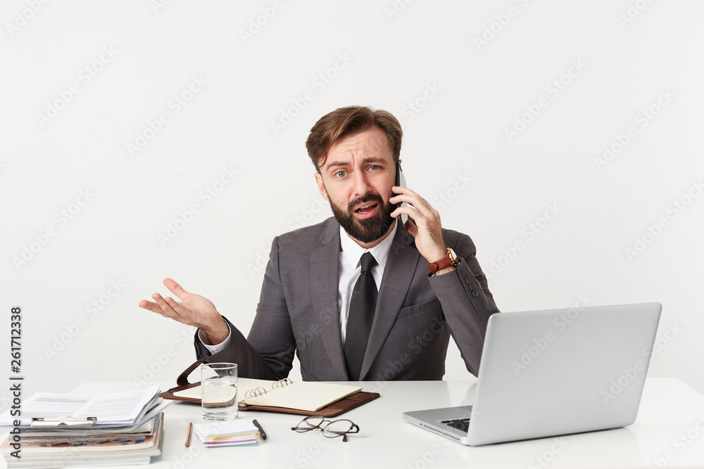 Shot of respectable office worker sitting at desktop, has unpleasant telephone conversation, feels unhappy cheated, he is told ridiculous things, dressed in a suit with a tie, has beard Shot of respectable office worker sitting at desktop, has unpleasant telephone conversation, feels unhappy cheated, he is told ridiculous things, dressed in a suit with a tie, has beard