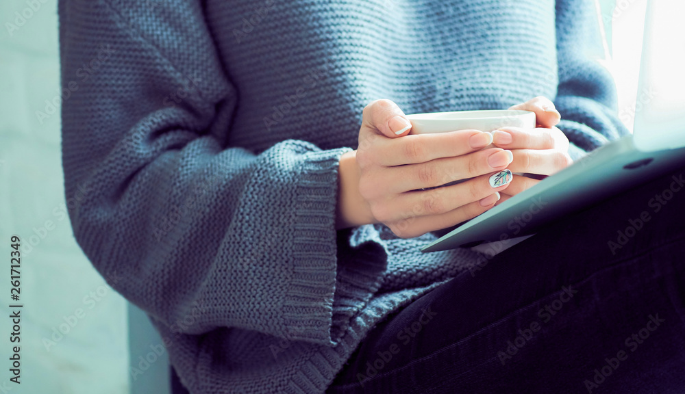 Girl sending a message, e-mail, woman typing something on the laptop in her lap sitting near window and holding cup of coffee close-up. Girl sending a message, e-mail, woman typing something on the laptop in her lap sitting near window and holding cup of coffee close-up.