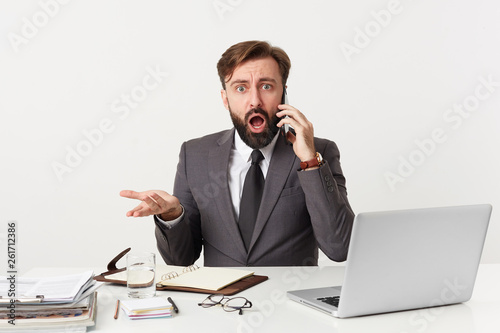 Close up of dressed in a suit respectable office worker sitting at desktop looking straight in confusion disbelief, stunned holding a phone near his ear, mouth opened from shocking news.