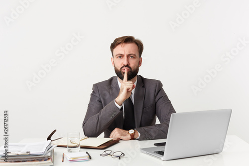 Serious volitional calm emotionless bearded man in business suit, showing silence gesture, asks to keep secret privacy stay quiet putting fore finger on the lips, isolated over white background