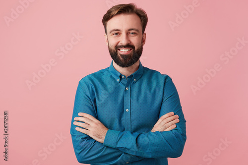 Representative, personable man with beard looking camera with pleasant smile, stands with arms folded, isolated over pink background, dressed in a blue shirt.