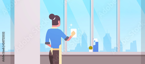 woman in gloves and apron cleaning windows with rag cleaner spray rear view housewife doing housework concept modern living room interior flat horizontal potrait