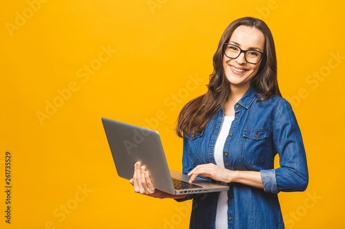 Young happy smiling woman in casual clothes holding laptop and sending email to her best friend. Isolated against yellow background.