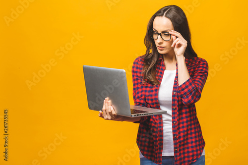 Young happy smiling woman in casual clothes holding laptop and sending email to her best friend. Isolated against yellow background.