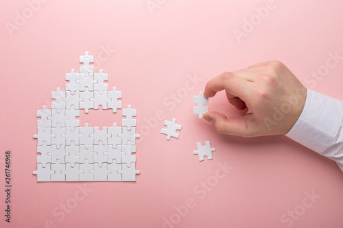 White puzzle. House shape puzzle. The concept of rent, mortgage. Hand holding piece of white puzzle. Pink background. photo