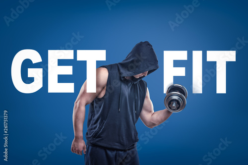 Athletic man in sleeveless hoody posing half-turn with dumbell in one hand on blue background with title GET FIT. photo