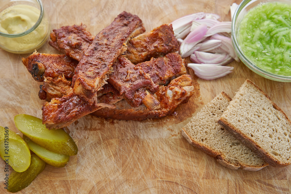 Top view on spicy bbq spare ribs marinated and served on wooden plate with slices of rustic bread, pickled cucumbers, french style mustard, chopped onion and fresh cucumber salad in glass bowl. 