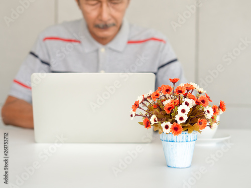 Blurred images of Asian elderly men enjoying the use of laptops on the white table, He is studying the use of modern technology..Focus on fake flowers in light blue pot placed in front.