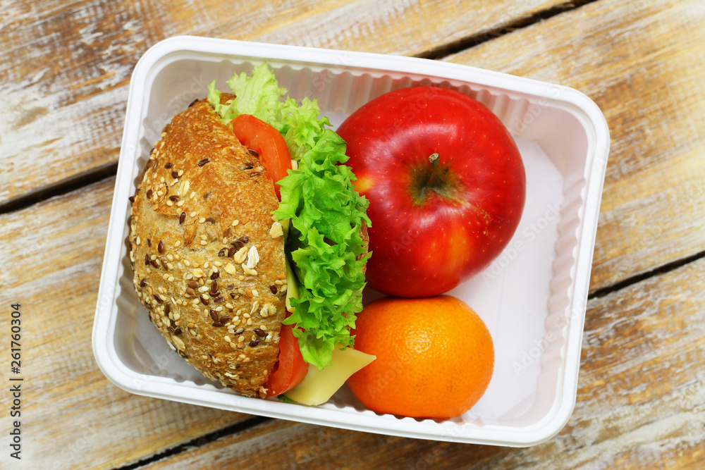 Healthy lunchbox containing brown cheese roll with lettuce and tomato, apple and orange