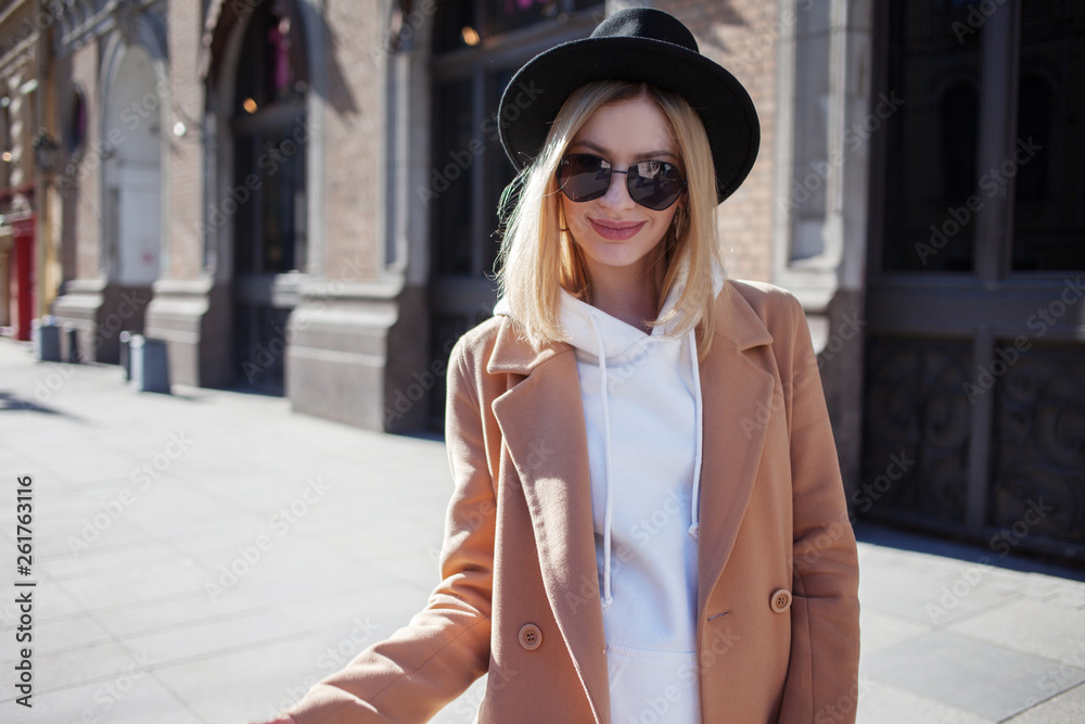 Fototapeta premium Happy girl outdoors, Sunny day. Fashionable and trendy young woman in black felt hat and stylish beige coat