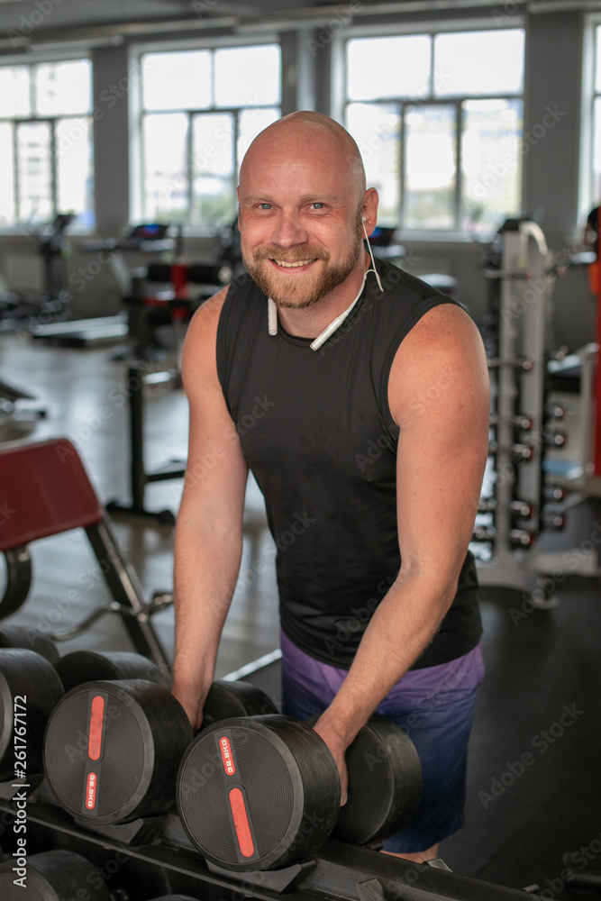 handsome young man in blue shorts and black t-shirts, doing exercises for biceps in the gym. bald man smiling at camera