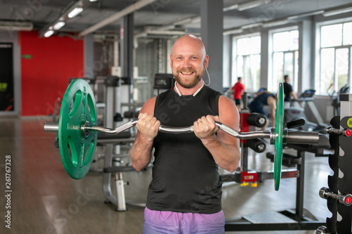 Bald athlete in a black T-shirt and blue shorts with headphones, lifting the barbell with effort. He is standing in the gym and smiling