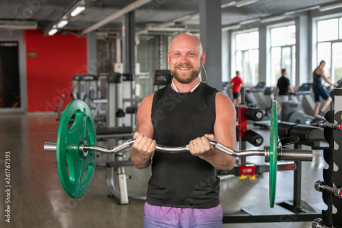 Bald athlete in a black T-shirt and blue shorts with headphones, lifting the barbell with effort. He is standing in the gym and smiling