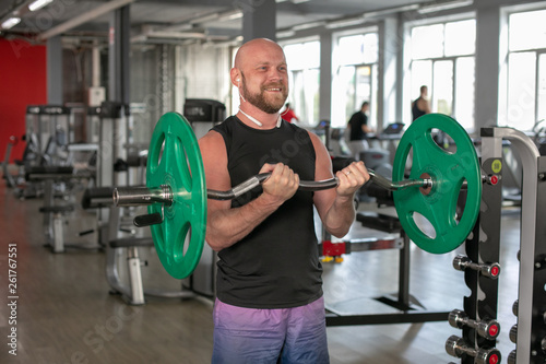 Bald athlete in a black T-shirt and blue shorts with headphones, lifting the barbell with effort. He is standing in the gym and smiling