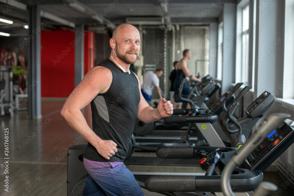 caucasian Man With Headphones Running on Treadmill in Gym. bald.man smiling at camera