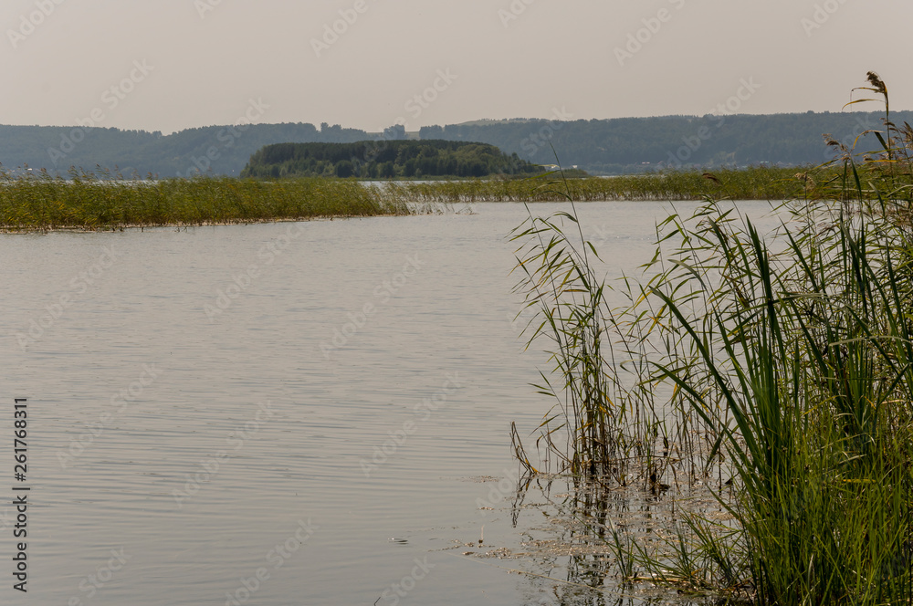 Wide blue river slow flowing across the green meadow and mountains with no reflections. A lot of tall grass around. Summer time Wide blue river slow flowing across the green meadow and mountains with no reflections. A lot of tall grass around. Summer time