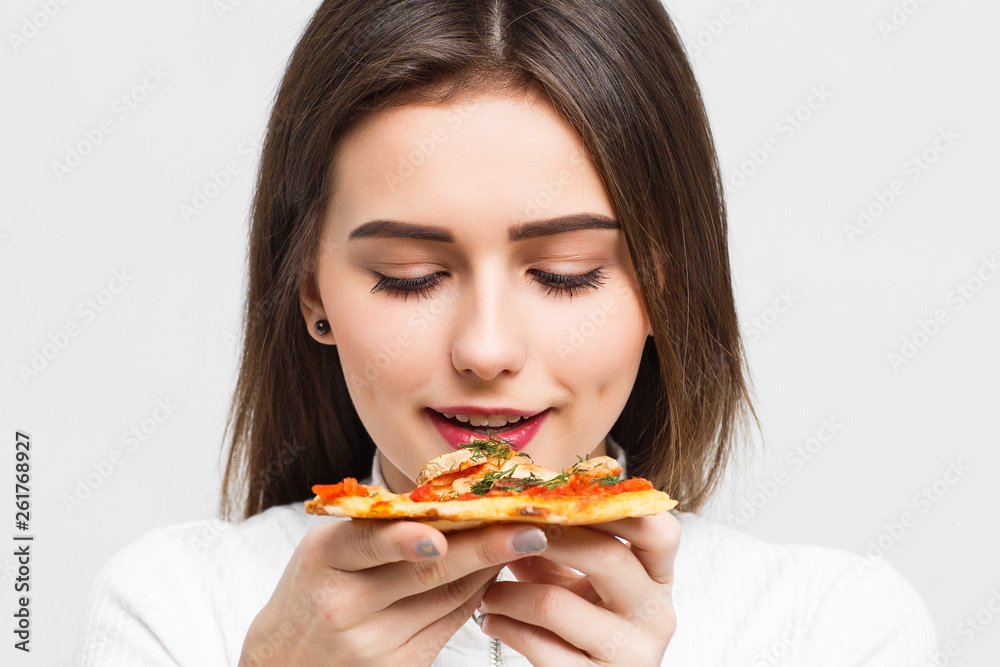 Mmm, so delicious! Dark haired pretty woman eats slice of Italian pizza, keeps eyes closed from pleasure, enjoys nice taste, isolated over white background.
