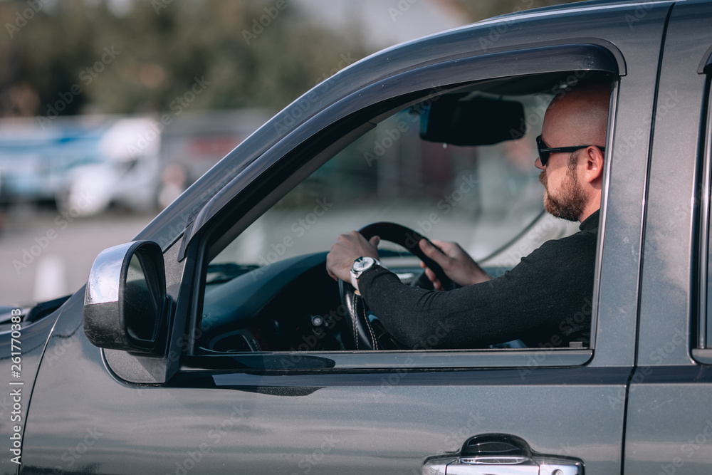 bald and bearded male in glasses with a clock in a suit behind the wheel of a car