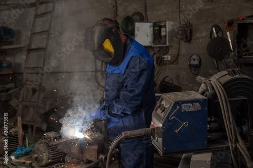  professional welder is welding a metal construction in garage wearing mask, proctive glasses and blue uniform. Blue sparks are flying apart.