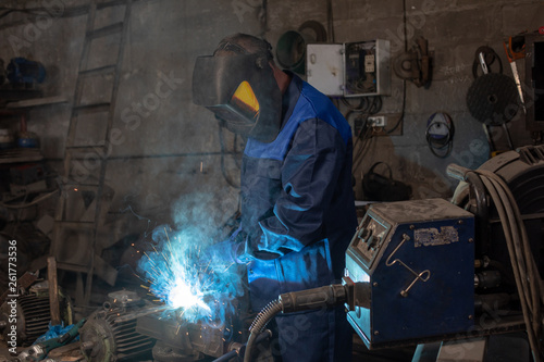 Strong professional welder is welding a metal construction in garage wearing mask, proctive glasses and blue uniform. Blue sparks are flying apart.