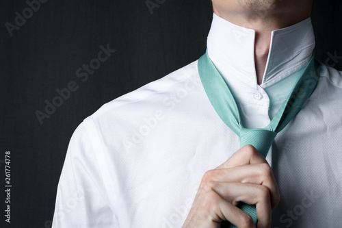 Close-up young man in a white shirt with a tie color Mint. The man straightens his tie. Businessman in a white shirt and turquoise necktie. Dark background. Copy space. Neo Mint color of the year 2020