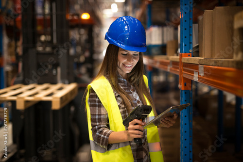 Smiling female worker holding tablet and bar code scanner checking inventory in distribution warehouse. Forklift in background.