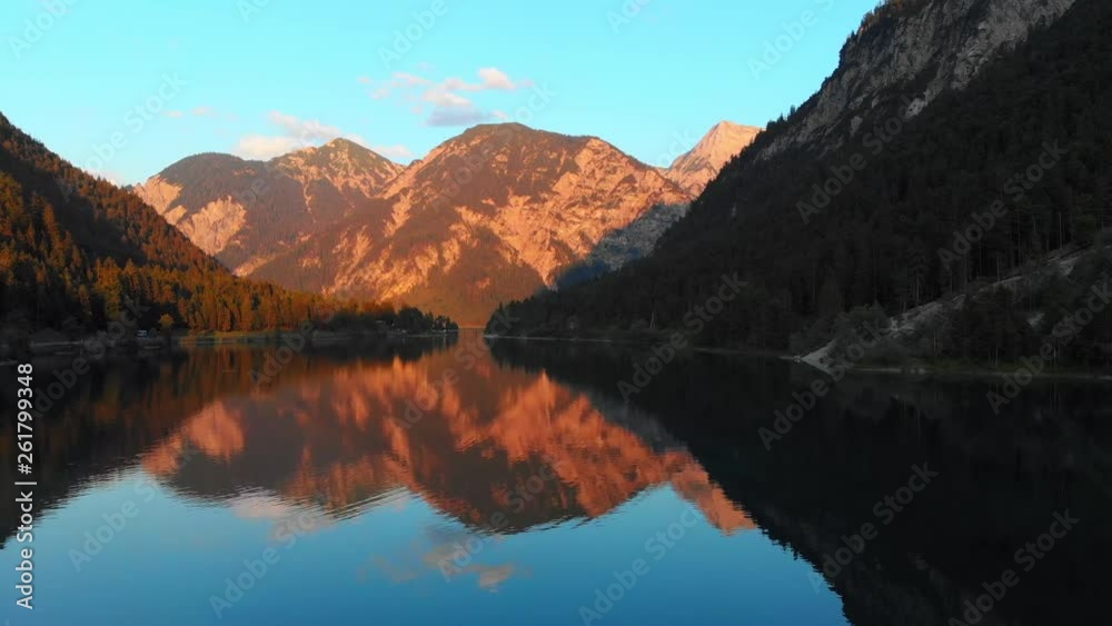 Aerial, drone shot, bypassing a spruce tree, towards sunset colored mountains, above reflecting waters, on lake Plansee, on a sunny summer evening, in Tyrol, Austria Aerial, drone shot, bypassing a spruce tree, towards sunset colored mountains, above reflecting waters, on lake Plansee, on a sunny summer evening, in Tyrol, Austria