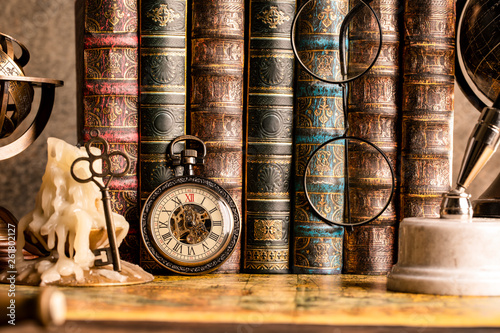 Antique clock on the background of vintage books. Mechanical clockwork on a chain. Fountain pen and glasses.
