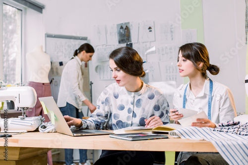 Two attractive women dressmakers designers choosing trendy sketches and materials for new collection of clothes, while young girl designer working on new model tailoring pants on mannequin in studio