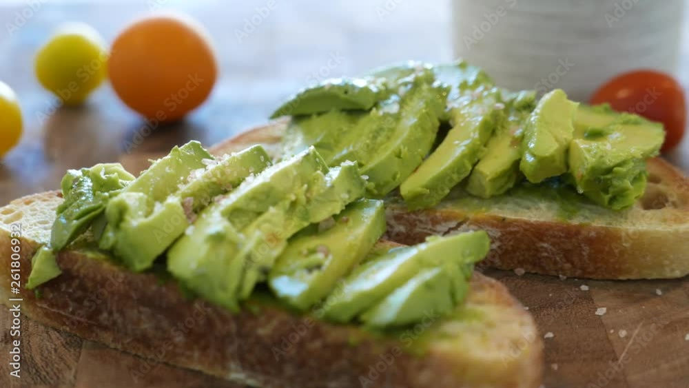 Rustic avocado toast sits on an artisan cutting board with salt falling slowly.
