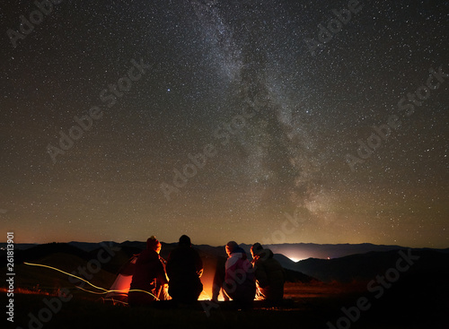 Back view group of four young friends hikers resting together around bonfire near camp and tourist tent at night in the mountains. On background beautiful night starry sky full of stars and Milky way.