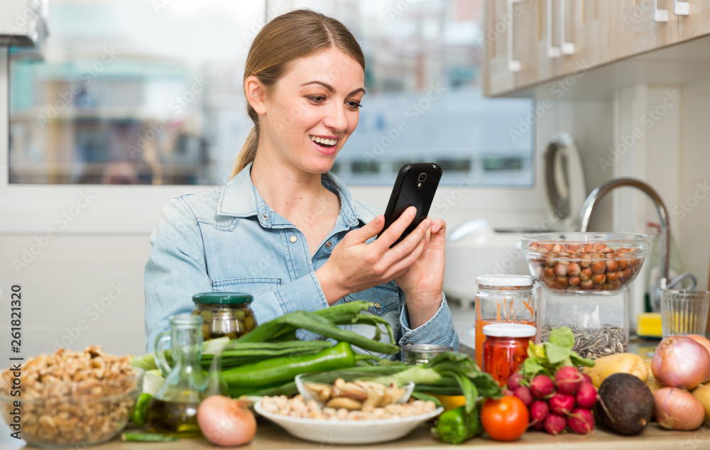 Portrait of glad woman looking at mobile phone while making dish on kitchen at home