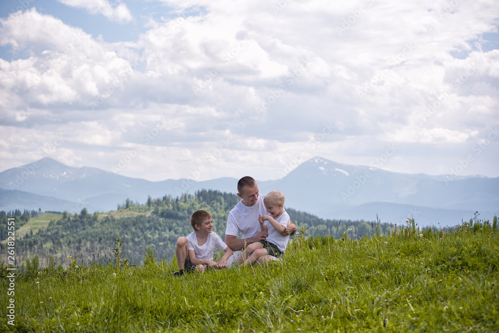 Happy father with his two young sons sitting on the grass on a background of green forest, mountains and sky with clouds. Friendship concept. Happy father with his two young sons sitting on the grass on a background of green forest, mountains and sky with clouds. Friendship concept.